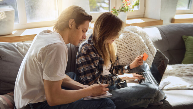 Couple comparing online payment options with laptop and bank card on sunny sofa. Concept of home ecommerce, secure checkout, shared expenses and digital banking.