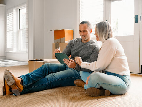 Happy couple seated on carpeted floor of their new home holding a tablet device