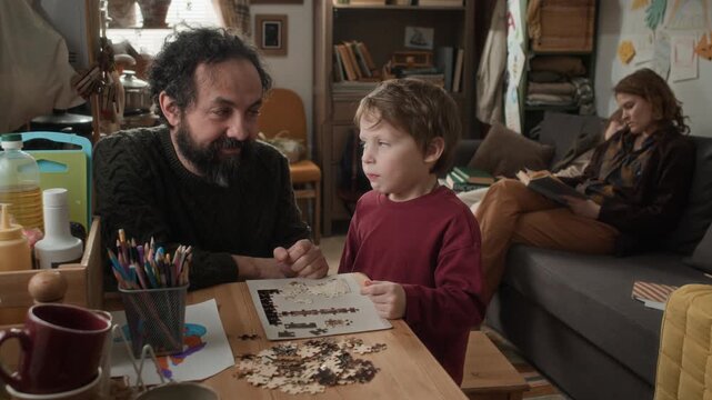 Portrait shot of father and son looking at camera while putting together puzzle at table while mother and brother napping on couch in small poor apartment