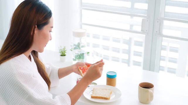 Young woman in bathrobe preparing breakfast putting jam on bread .