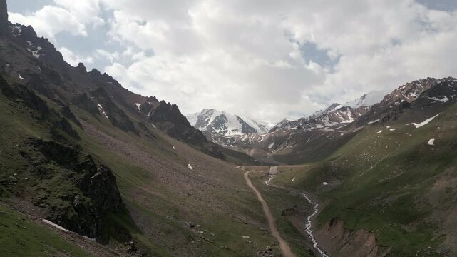 Winding dirt road in a deep mountain valley with snow peaks, aerial view.