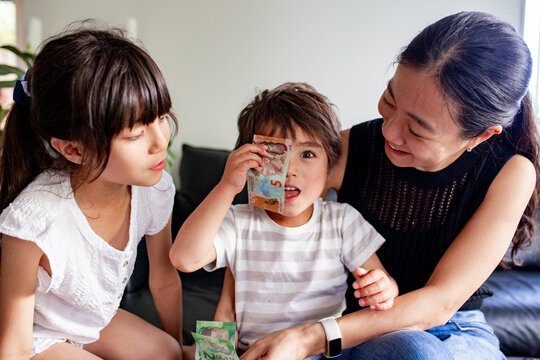 Toddler sitting between mum and older sister covering one eye with five NZ dollar bill in the couch