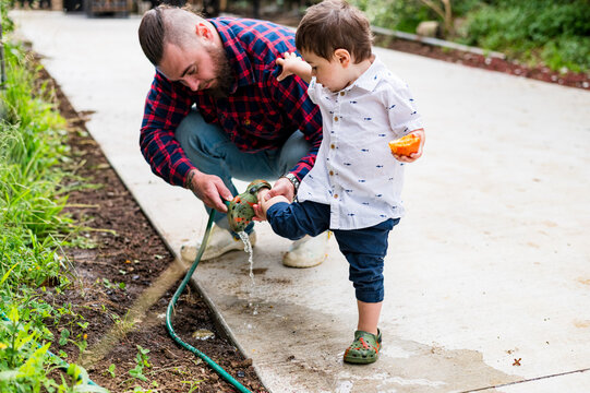 Father in plaid shirt crouches on a paved garden pathway washing the dirt off toddler sons shoes
