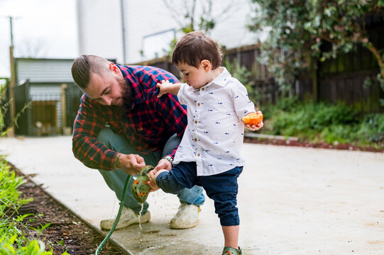 Father in plaid shirt crouches on a paved garden pathway washing the dirt off toddler sons shoes