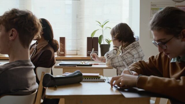 Medium side shot of diverse group of high school students writing test at lesson, geeky Caucasian teenage boy in glasses thinking then choosing correct answers on exam paper