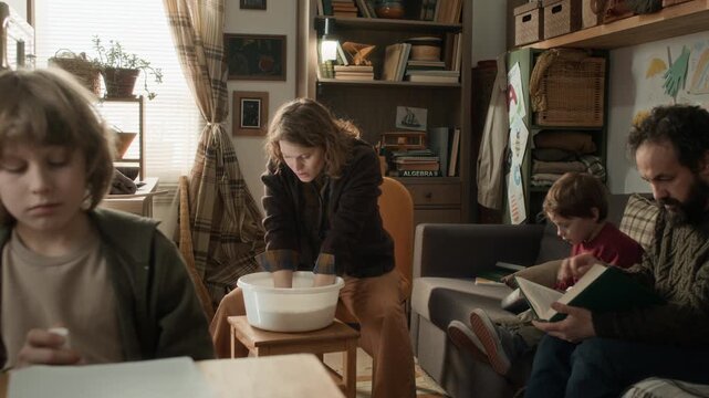 Mom doing laundry by hand, dad and younger son reading book on couch, and older son sitting at table in cramped but cozy room