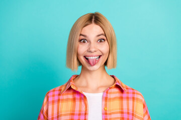 Fototapeta premium Playful young woman with blonde bob sticking out her tongue in a colorful plaid shirt on turquoise background, cheerful portrait expressing fun and carefree attitude
