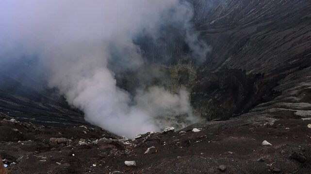 Inside Mount Bromo volcano crater with dense volcanic steam rising from active vents. Dark ash-covered crater walls and rugged volcanic terrain in East Java, Indonesia.