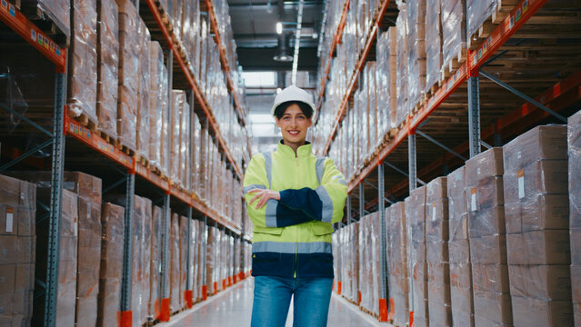 Caucasian female warehouse worker standing confidently with arms crossed, wearing safety jacket and helmet. Positioned in central aisle surrounded by stacked boxes on high industrial shelves.