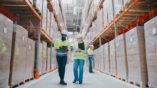 Warehouse managers Man holding tablet, woman with clipboard, communicating during inspection. Other people passing by.