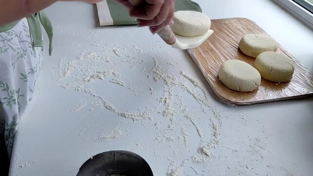 Hands of a female chef preparing homemade syrniki (cottage cheese pancakes) by coating them in flour on a wooden board before frying. Home cooking, traditional recipes, and culinary preparation.