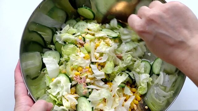 In the close-up image, a woman's hands are mixing fresh salad in a stainless steel bowl. Bright ingredients - sliced cucumbers, iceberg lettuce, tuna, eggs, celery, corn