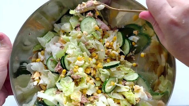 In the close-up image, a woman's hands are mixing fresh salad in a stainless steel bowl. Bright ingredients - sliced cucumbers, iceberg lettuce, tuna, eggs, celery, corn