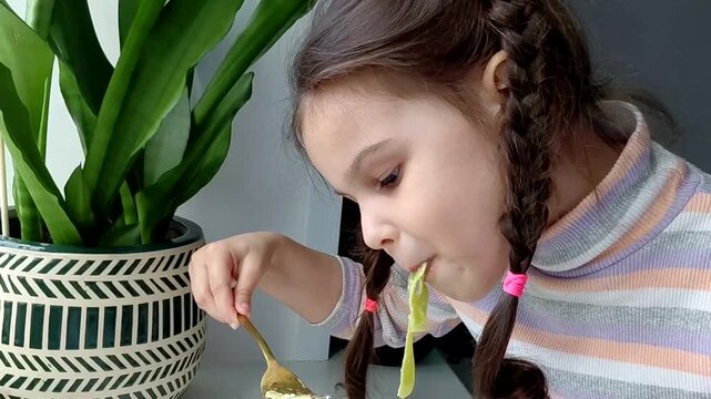 A little girl eats pasta with vegetables near the window. Funny child eats vegetables and laughs