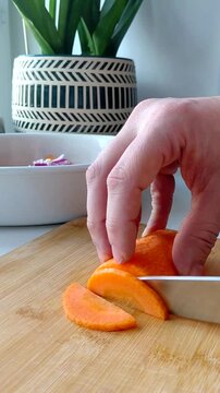 A person slices vibrant orange carrots with a knife on a rustic wooden cutting board