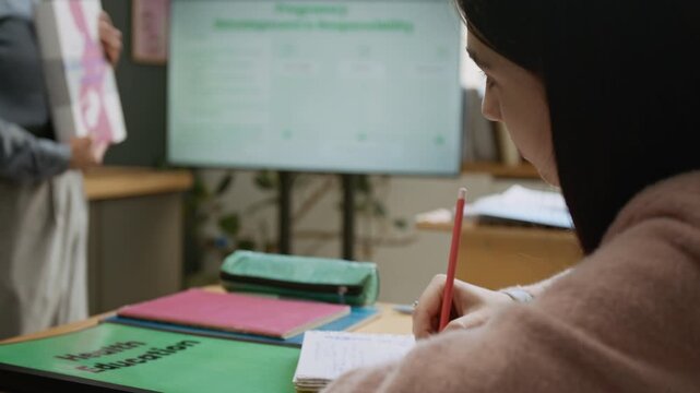 Medium close-up of diligent young girl taking notes in copybook during health education lesson at high school, female teacher with pregnancy textbook talking to students by blackboard