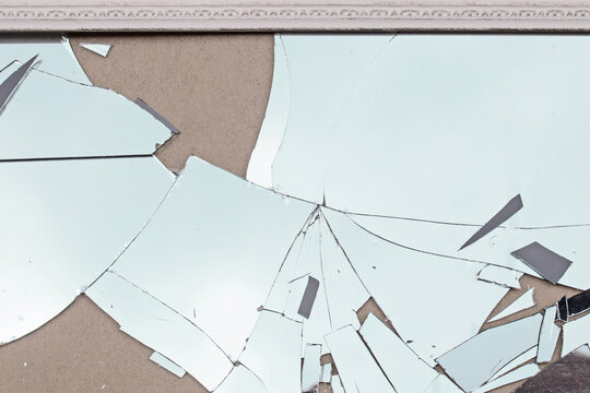Broken mirror glass on a wall with scattered shards and a frame at a home setting in daylight hours