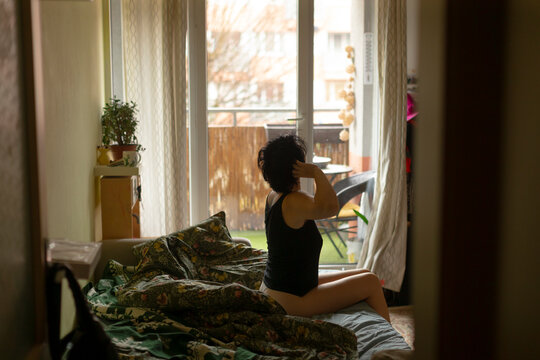asian woman dressing by window, dancer adjusting undergarment before rehearsal, warm light, patterned quilt pooled on mattress, candid selfcare moment with plants and open balcony.