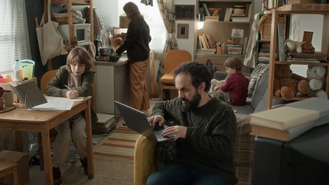 Man working on his laptop, boy doing his homework, his brother playing on couch, and woman cooking in kitchen, in cramped, run-down apartment