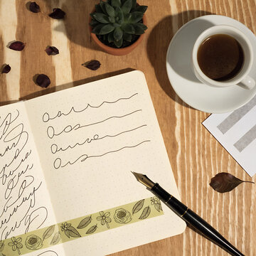 Open notebook showing cursive letter practice lines decorated with decorative washi tape alongside a classic fountain pen and a cup of dark coffee.