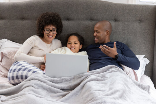 Diverse family sitting on bed in bedroom, child holding silver laptop under plush gray blanket