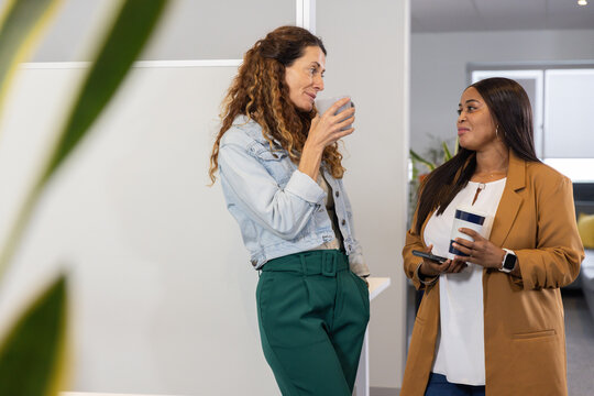 Diverse women staff standing, chatting in break area, holding mug and travel cup, denim, tan blazer