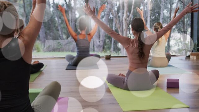 Women practicing yoga following cue, sweeping arms upward on mats by glass wall, bokeh drifting
