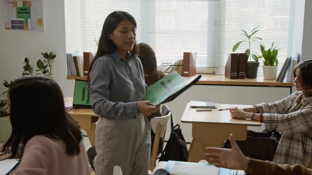 Medium tracking shot of young female teacher walking around classroom, giving diverse teenage students folders with lesson material handouts and test questions on health education