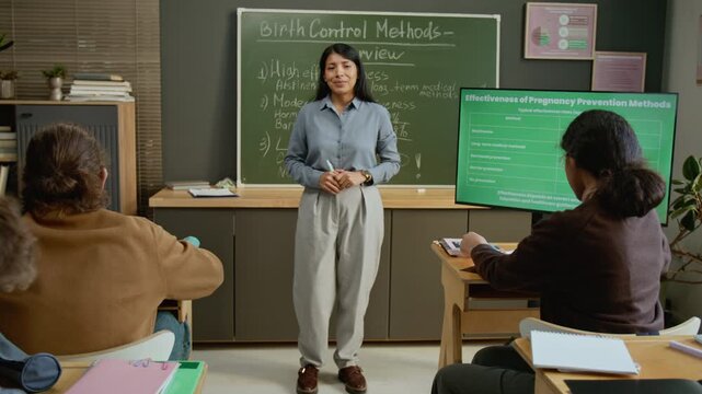 Full dolly portrait of smiling young Hispanic female high school teacher standing in front of blackboard at reproductive health lesson, looking at camera while students writing test on birth control
