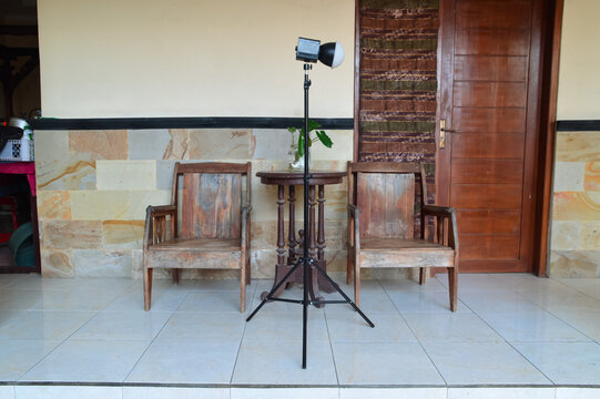 A professional black photography light with a softbox is mounted on a tall tripod stand on a tiled house porch, positioned in front of wooden chairs and a small table for an outdoor photoshoot.