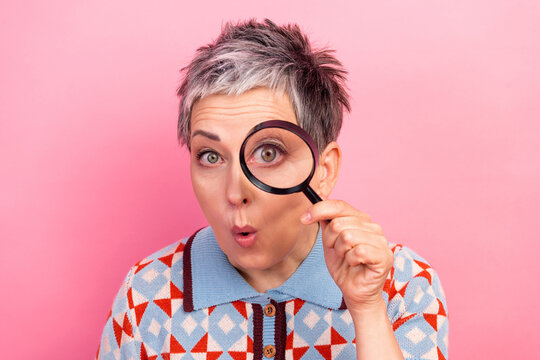 Surprised woman with magnifying glass examining camera on pink background, playful portrait of curious middle-aged woman with short gray hair and patterned sweater