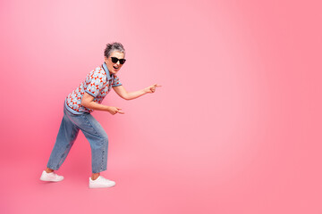 Obraz premium Happy stylish senior woman in sunglasses pointing and laughing against a pink background, casual retro outfit and confident playful pose