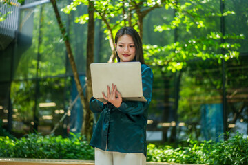 Naklejka premium Professional young Asian woman using a laptop in a lush outdoor park with a modern glass building background. Ideal for remote work, technology, education, and digital nomad lifestyle themes.
