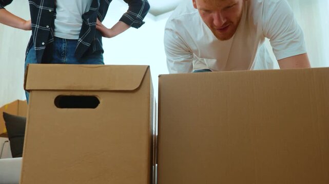 Close up of positive young man and woman in room with many boxes putting stuff on floor. Married couple with cardboard boxes indoor in new apartment. Moving day concept. Real time