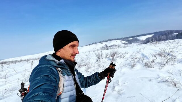 Caucasian Man Trekking Across Snowy Plain With Orange Backpack And Ski Poles, Wearing Blue Jacket And Black Beanie, Panoramic Winter Landscape Under Clear Sky, Crisp Daylight And Solitary