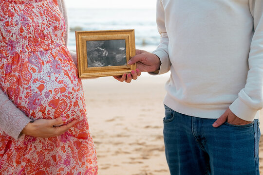 Pregnant woman  with husband on sand near ocean. Mother and father hugging in anticipation of baby and holding photo of baby's girth in golden wooden frame. Pregnancy planning in sunny Portugal
