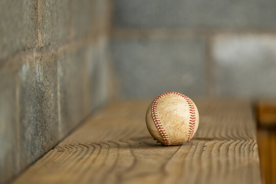 Worn baseball sits on a wooden dugout bench in close-up, a weathered cinderblock wall filling the left side of the frame in a textural baseball detail image.