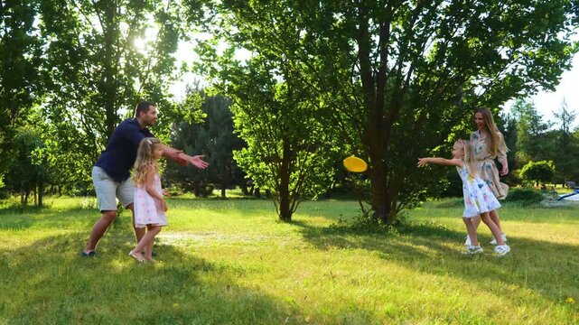 Positive family throwing frisbee disk in park. Smiling girl catching frisbee plate. Young parents playing frisbee with daughters. Family enjoying summer outdoor activity. Real time. Happiness