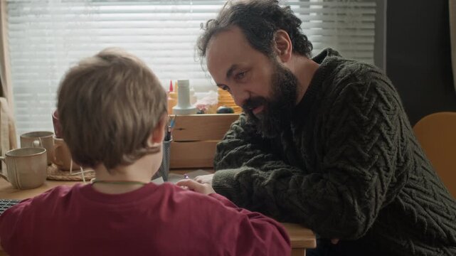 Close up shot of bearded man coloring with his young son and patting him by shoulder at wooden table