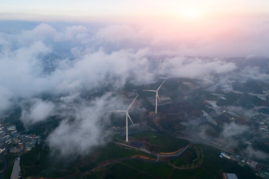 White turbines pierce morning clouds, blades spinning for energy. Sunlight bathes the misty, green valley below