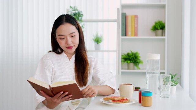 Smiling young asian woman having breakfast, coffee and bread  while enjoy reading book . morning routine.