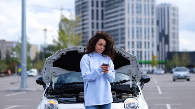 Portrait of beautiful young woman standing outdoor near broken car waiting for help evacuator. Female in street near vehicle with opened hood. Car breakdown. Car accident. Real time video