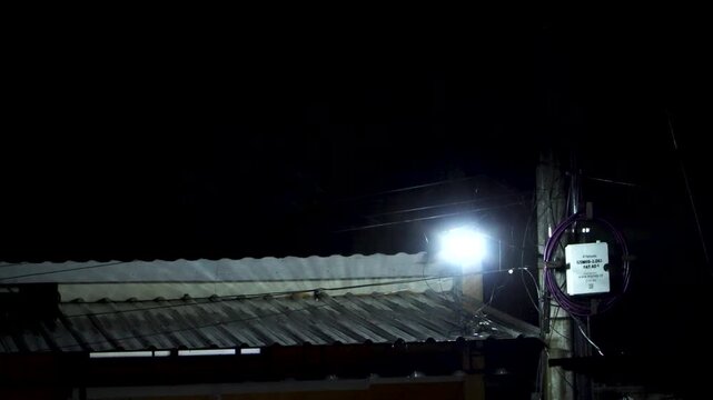 Bright street light and utility pole with a Wi-Fi cable box illuminating a residential tin roof during a dark rainy night in East Java, Indonesia.