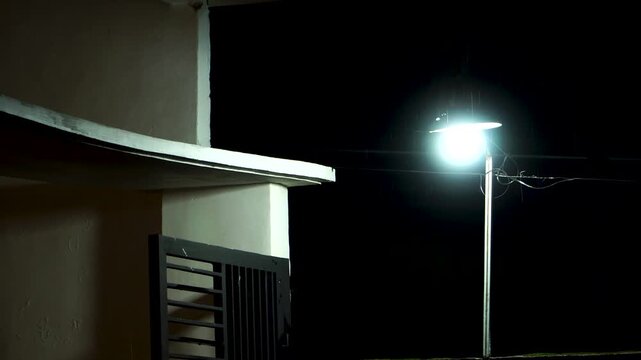 Handheld shot of a bright outdoor light illuminating falling rain by a house terrace and black gate during a dark, stormy night in an Indonesian residential neighborhood.