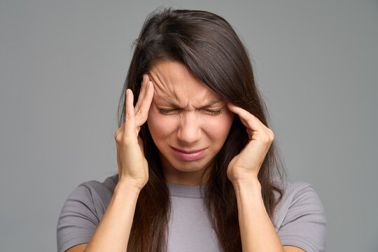 Tight portrait of woman with clenched eyes pressing temples, neutral grey studio