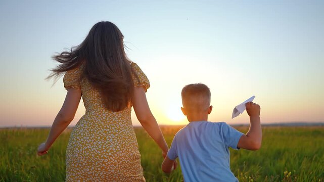 Mother and son run in green field at sunset holding hands. Happy boy holds paper airplane while running with woman. Family enjoys summer sunset outdoors. Mother and son play with paper airplane.
