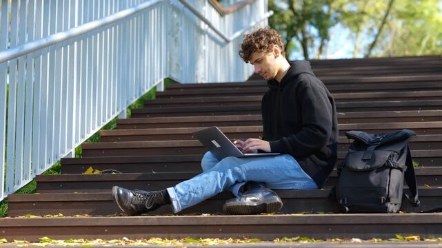Young man sitting on the staircase in the city park while using laptop and typing. Autumn season. Education, working concept. Real time concept.