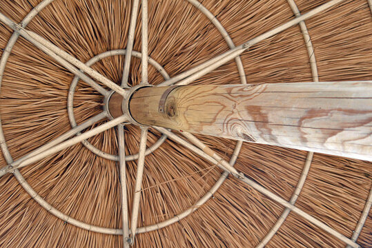 Nahaufnahme eines h&ouml;lzernen Sonnenschirms mit traditionellem Strohdach, dessen Gestell aus radialem Bambus und Naturholzbalken besteht. Tropisches Stranddesign mit rustikaler Handwerkskunst, starker S