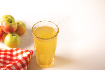 Fototapeta premium A refreshing glass of golden apple juice or cider with a reusable straw, surrounded by fresh apples and a red checkered cloth on a light background.
