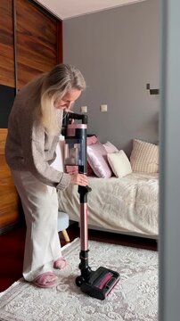 Blonde woman folds cordless vacuum cleaner after finishing bedroom cleaning and leaves room. End of household routine, tidy home satisfaction and organized lifestyle concept.
